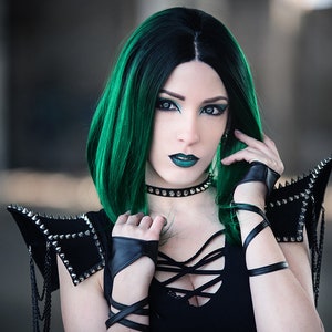 May include: A woman with green hair and dark makeup is wearing a black top with a lace-up front and black leather gloves. She is also wearing a black choker with silver studs and a pair of black leather shoulder pads with silver studs.
