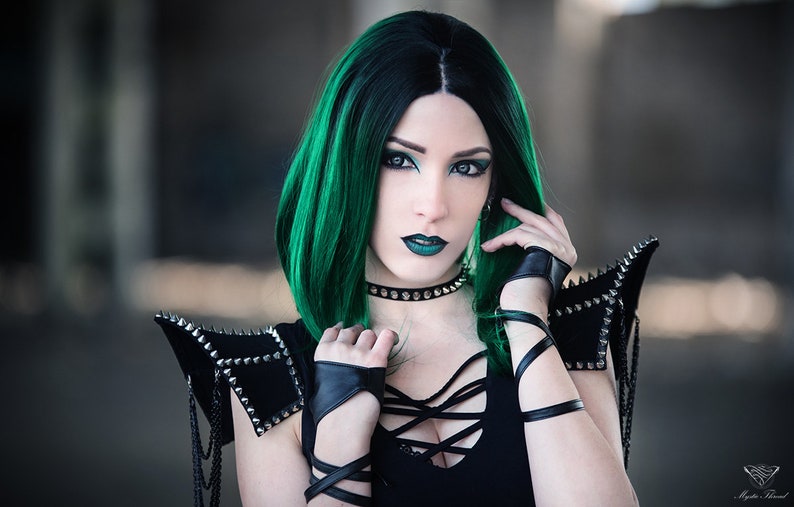 May include: A woman with green hair and dark makeup is wearing a black top with a lace-up front and black leather gloves. She is also wearing a black choker with silver studs and a pair of black leather shoulder pads with silver studs.