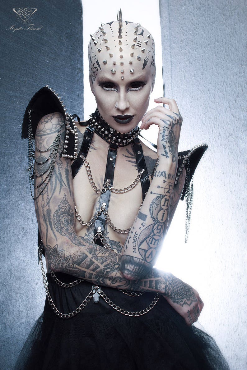May include: A person with a shaved head covered in silver studs and black ink tattoos is wearing a black leather harness with silver chains and a black choker with silver studs. The person is wearing a black tulle skirt with silver chains.