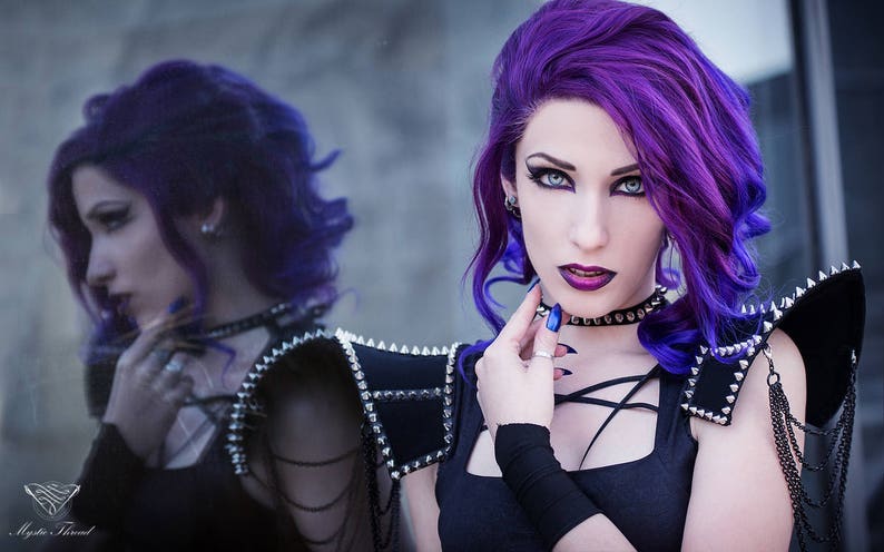 May include: A woman with long, bright purple hair wears a black top with a criss-cross design and a studded black leather shoulder piece. She is wearing a black choker with silver studs and has dark eye makeup.