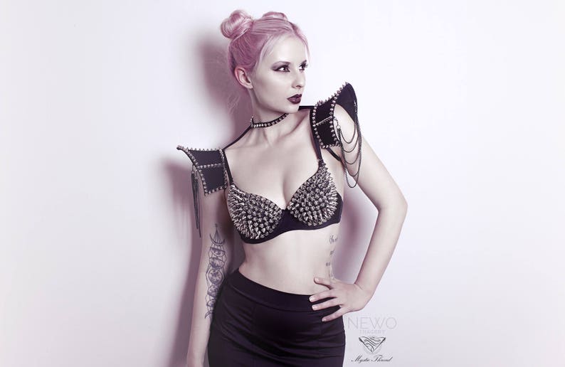 May include: A woman with pink hair is wearing a black bra with silver studs and a matching black studded shoulder piece. The shoulder piece has silver chains hanging from it.