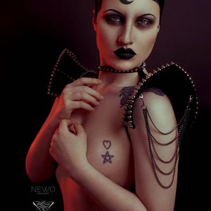May include: A person with black hair and dark makeup is wearing a black leather shoulder piece with silver studs and chains. The shoulder piece has a pointed design and is worn on both shoulders. The person is also wearing a black choker with silver studs. The person has a tattoo of a heart and a star on their chest.