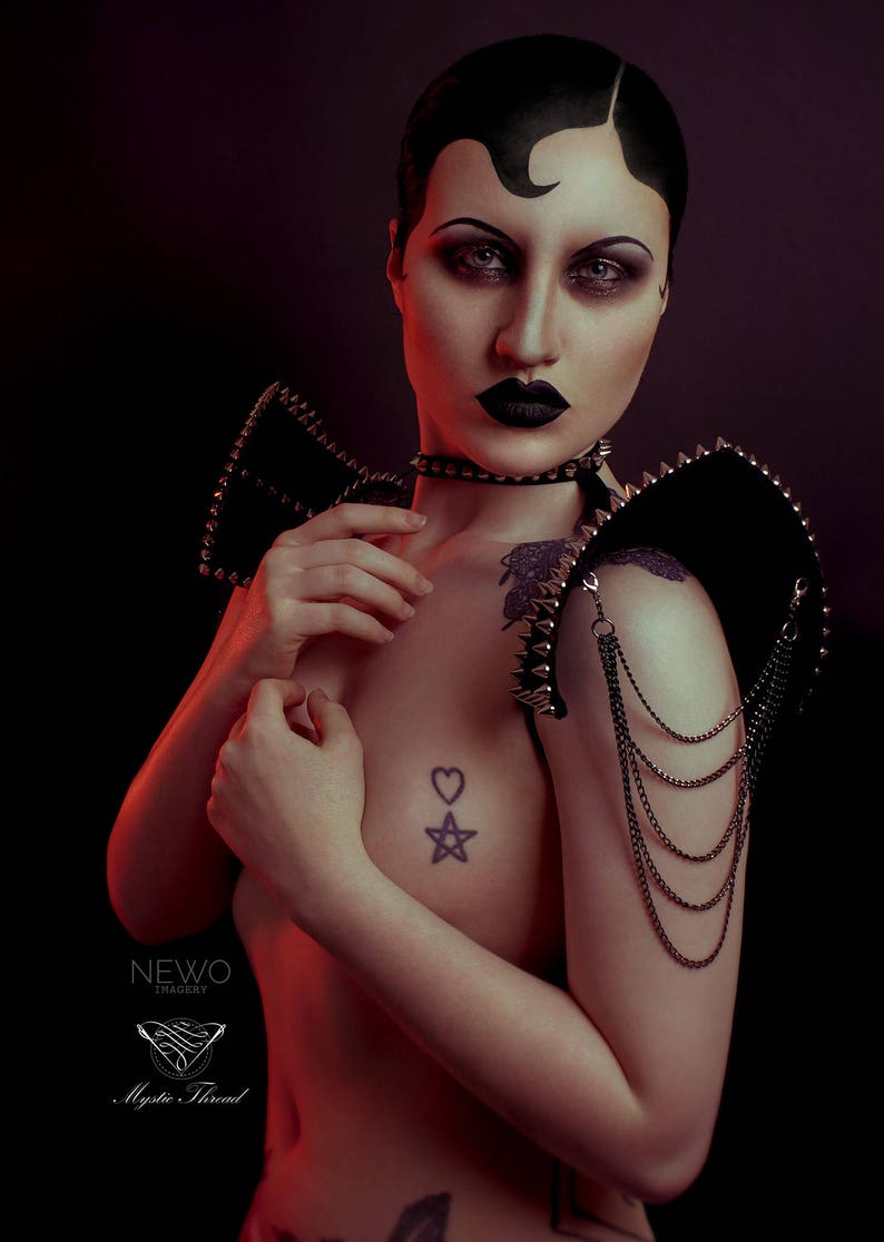 May include: A person with black hair and dark makeup is wearing a black leather shoulder piece with silver studs and chains. The shoulder piece has a pointed design and is worn on both shoulders. The person is also wearing a black choker with silver studs. The person has a tattoo of a heart and a star on their chest.