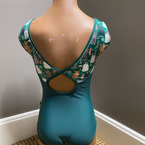 May include: A dark green leotard with a low back and a keyhole cutout. The leotard has short sleeves with a festive print of cats and Christmas decorations.