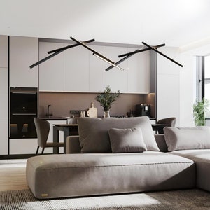 Op de afbeelding: Een moderne woonkamer met een grote hoekbank in een lichte grijze stof. De bank heeft meerdere kussens en staat voor een groot raam. De kamer heeft een moderne keuken met witte kasten en een zwarte oven. Een zwarte lineaire kroonluchter hangt boven de eettafel.