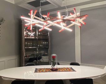 May include: A modern dining room featuring a large white oval table surrounded by black and white chairs. A unique, abstract chandelier with red and white lights hangs above the table. A wine rack and a glass cabinet are visible in the background.