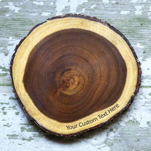 May include: A round wooden slice with a natural wood grain pattern. The slice has a light brown outer ring and a darker brown center. The text "Your Custom Text Here" is engraved on the outer ring.