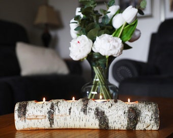 May include: A white birch log candle holder with five candle openings. The log is sitting on a wooden table with a bouquet of white flowers in a glass vase in the background.