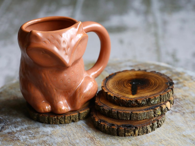 May include: A ceramic fox mug with a brown glaze sits on a set of three wooden coasters made from tree slices.