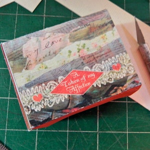 Op de afbeelding: Een kleine doos met rode randen en een collage-ontwerp. De bovenkant heeft gelaagde papieren stroken met bloemen- en scriptpatronen. Een rode ruit met de tekst "A Token of my Affection" is gecentreerd, met kant- en hartaccenten.