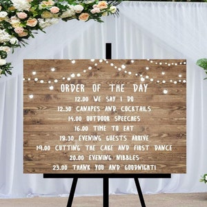 Peut inclure: Panneau de mariage en bois sur un chevalet noir, affichant l'&laquo;&nbsp;Ordre du jour&nbsp;&raquo; en texte blanc. Le panneau est d&eacute;cor&eacute; de guirlandes lumineuses et entour&eacute; de compositions florales. Le texte comprend le programme du mariage, tel que &laquo;&nbsp;We Say I Do&nbsp;&raquo; et &laquo;&nbsp;Cutting the Cake&nbsp;&raquo;.