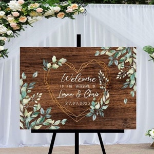 May include: A rustic wooden wedding welcome sign with a gold geometric heart frame and green watercolor foliage. The sign reads "Welcome to the wedding of Lorna & Chris 27.07.2023".