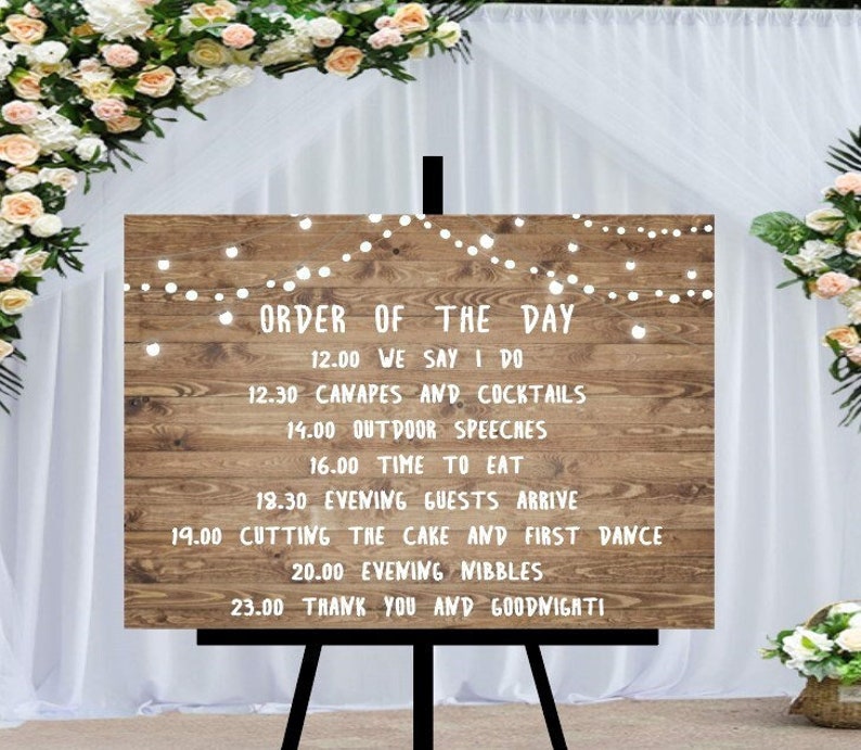 Peut inclure: Panneau de mariage en bois sur un chevalet noir, affichant l'&laquo;&nbsp;Ordre du jour&nbsp;&raquo; en texte blanc. Le panneau est d&eacute;cor&eacute; de guirlandes lumineuses et entour&eacute; de compositions florales. Le texte comprend le programme du mariage, tel que &laquo;&nbsp;We Say I Do&nbsp;&raquo; et &laquo;&nbsp;Cutting the Cake&nbsp;&raquo;.