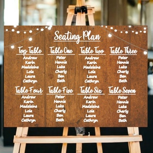 May include: A rustic wooden seating chart with white text and fairy lights. The chart lists the names of guests assigned to each table, including "Top Table", "Table One", "Table Two", "Table Three", "Table Four", "Table Five", "Table Six", and "Table Seven".