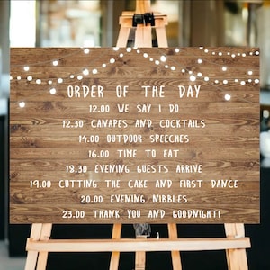 Peut inclure: Panneau en bois sur chevalet affichant l'&laquo;&nbsp;Ordre du jour&nbsp;&raquo; d'un mariage. Le panneau pr&eacute;sente un fond en bois rustique avec du texte blanc et des guirlandes lumineuses d&eacute;coratives. L'horaire comprend les heures de la c&eacute;r&eacute;monie, des discours, du d&icirc;ner et de la danse.