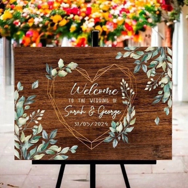 May include: A wooden welcome sign with a gold geometric heart design and green watercolor leaves. The sign reads "Welcome to the wedding of Sarah & George 31/05/2024".
