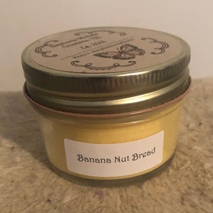 May include: A small jar candle with a metal lid. The candle has a yellow wax and a label that reads "Banana Nut Bread." The lid has a decorative label with text and a butterfly illustration. The jar sits on a textured surface.