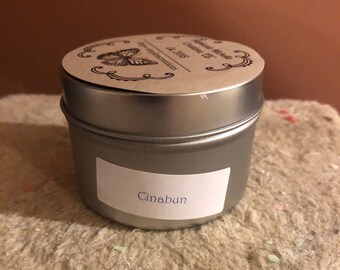 May include: A silver metal tin candle with a brown paper label that says "Cinnabun" and a butterfly design on the lid. The label also says "Handmade Candles" and "Est. 2015".