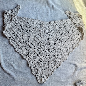 May include: A crocheted shawl in a cream color with a scalloped edge. The shawl is made with a delicate stitch pattern and has a triangular shape.