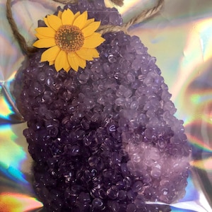 May include: A purple and light purple car air freshener shaped like a heart with a yellow sunflower on top. The air freshener is tied with a brown twine.