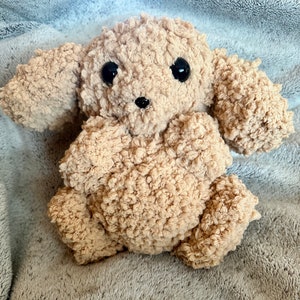May include: A soft, plush, beige-colored stuffed toy dog with black eyes and a black nose. The toy is sitting on a gray fuzzy blanket.