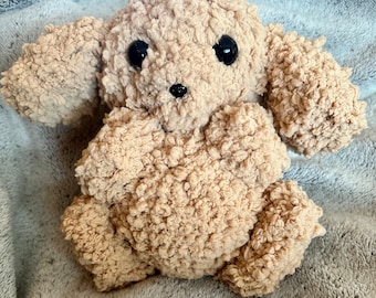 May include: A soft, plush, beige-colored stuffed toy dog with black eyes and a black nose. The toy is sitting on a gray fuzzy blanket.