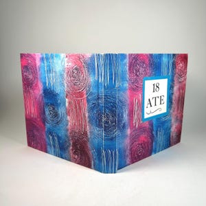 May include: A decorative box with a vibrant abstract design in shades of blue, pink, and white. The box features a textured pattern of swirls and vertical lines. The text "18 ATE" is displayed on a white label.