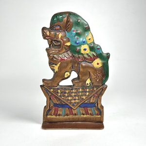 May include: A ceramic Foo Dog figurine, featuring a brown body with green, blue, and yellow accents. The mythical creature is posed on a decorative base with colorful geometric patterns. The figurine has an open mouth and detailed features.