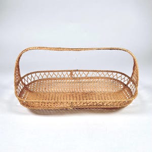 May include: A rectangular woven basket with a curved handle. The basket is made of light brown wicker and features an open weave design. The handle arches over the top, connecting to the basket's sides. The basket is empty and sits on a white surface.