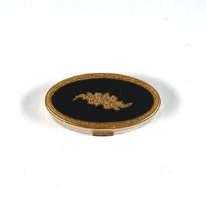 May include: An oval-shaped compact case featuring a black center inlay decorated with a gold-toned floral engraving. The perimeter of the lid is framed by an ornate, textured gold-colored metal border with a small latch visible at the front edge.