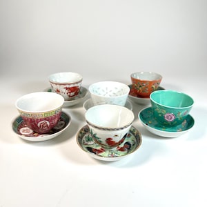 May include: A collection of six porcelain teacups and saucers. The cups feature various designs, including floral patterns, roosters, and Chinese characters. The saucers are decorated with complementary patterns and colors, such as red, green, and gold.