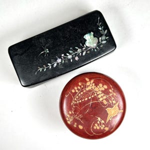 May include: A rectangular black box with mother-of-pearl floral and dragonfly inlay alongside a circular red box featuring a gold-toned bird and foliage design. Both containers are smooth and finished with decorative surface details.