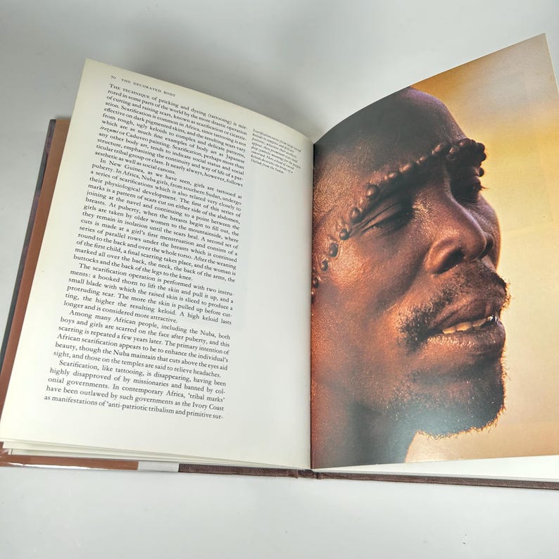 May include: Close-up portrait of a person with intricate scarification markings on their face. The markings are raised and create a textured pattern across the forehead, cheeks, and chin. The person's eyes are closed, and their mouth is slightly open, revealing a hint of a smile.