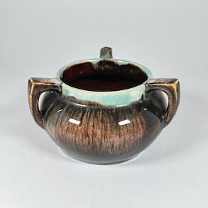 May include: A vintage ceramic sugar bowl with a brown and turquoise glaze. The bowl has two handles and a small handle on the lid. The interior is a dark brown color.