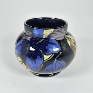 May include: A round, dark blue ceramic vase with a wide opening. The vase is decorated with large, stylized blue floral designs, with accents of yellow, pink, and brown. The vase has a glossy finish and sits on a white surface.