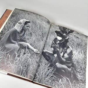 May include: A black and white photograph of two people in a grassy field. The person on the left is wearing a white head covering and a long, dark garment. The person on the right is wearing a headdress with feathers and a dark garment. The person on the right is holding a small object in their hand.