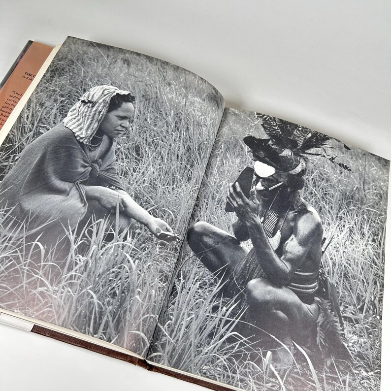 May include: A black and white photograph of two people in a grassy field. The person on the left is wearing a white head covering and a long, dark garment. The person on the right is wearing a headdress with feathers and a dark garment. The person on the right is holding a small object in their hand.