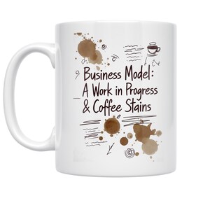 May include: White ceramic coffee mug with a handle. The mug features the text "Business Model: A Work in Progress & Coffee Stains" in black, along with brown coffee stain illustrations and doodles.