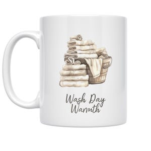 May include: White ceramic mug featuring a watercolor illustration of folded towels and a wooden bucket. The mug has the text "Wash Day Warmth" in a script font. The design is in shades of beige and brown.