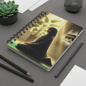 May include: A spiral-bound notebook featuring an illustration of a person in a black hijab kneeling in prayer. The artwork includes a large green potted plant and a depiction of the Kaaba in the background, set against a warm, golden light.