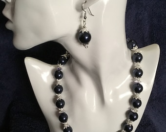 Puede incluir: Un collar y pendientes a juego de perlas negras. El collar está hecho de perlas negras con detalles plateados. Los pendientes son pendientes colgantes con una sola perla negra.
