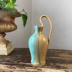 May include: A ceramic pitcher featuring a tall, curved handle and a two-tone glaze. One side of the vessel is a vibrant turquoise blue, while the other side is a warm, earthy tan, separated by a vertical band of muted sage green.