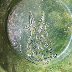 May include: A round, deep green ceramic bowl featuring a hand-etched, stylized line drawing of a horned creature with a long, curled tail. The surface has a glossy, mottled finish with visible texture and variations in the green glaze color.