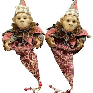 May include: Two identical monkey figurines dressed in jester-style outfits with pointed hats featuring a diamond pattern, ruffled collars, and long, tapered fabric tails in deep red with gold embroidery, accented by small bells and red beads.
