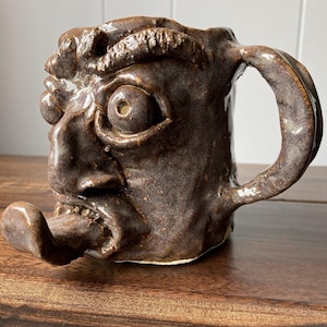 May include: A brown ceramic mug sculpted into the shape of a human face with a prominent nose, wide eyes, furrowed brows, and a protruding tongue extending from the mouth. The mug features a glossy, textured finish and a curved handle on the side.