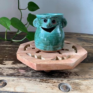 May include: A teal ceramic mug featuring a sculpted face with prominent ears, a nose, and a wide, open mouth. The mug has a glossy, variegated glaze finish with visible brush strokes and a smooth, rounded body shape, resting on a flat base.