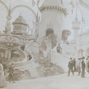 May include: A vintage sepia-toned photograph of an amusement park attraction featuring a large slide built into a rocky structure. The scene includes a tall tower with flags, arched walkways, and people dressed in early twentieth-century attire.