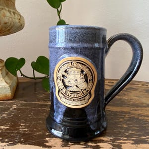 May include: A ceramic mug with a dark blue and black mottled glaze featuring a circular tan medallion on the front. The medallion displays the text Maryland Renaissance Festival Limited Edition and an illustration of a crown and goblet.