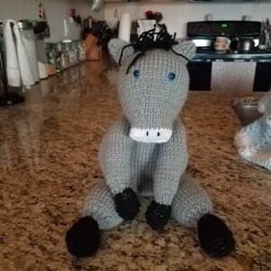 May include: A gray knitted horse toy with black hooves and blue eyes. The toy is sitting on a granite countertop.