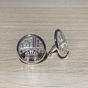 May include: A pair of silver cufflinks with a detailed cityscape design. The cufflinks are round and have a silver finish.